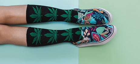 Two feet wearing colorful slip-on shoes and black and green cannabis socks