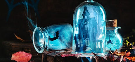 Three glass jars filled with gray smoke on a table: one with a bat, one with a haunted house, and one with a spider.