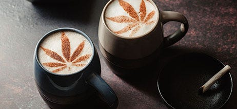 Two mugs of hot chocolate with cocoa sprinkled on top in the shape of cannabis leaves, sitting next to a joint in an ash tray