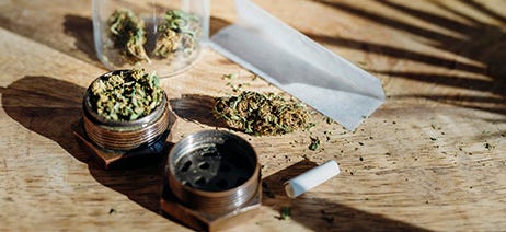 Cannabis flower in a mason jar and in a grinder, on a table next to rolling paper