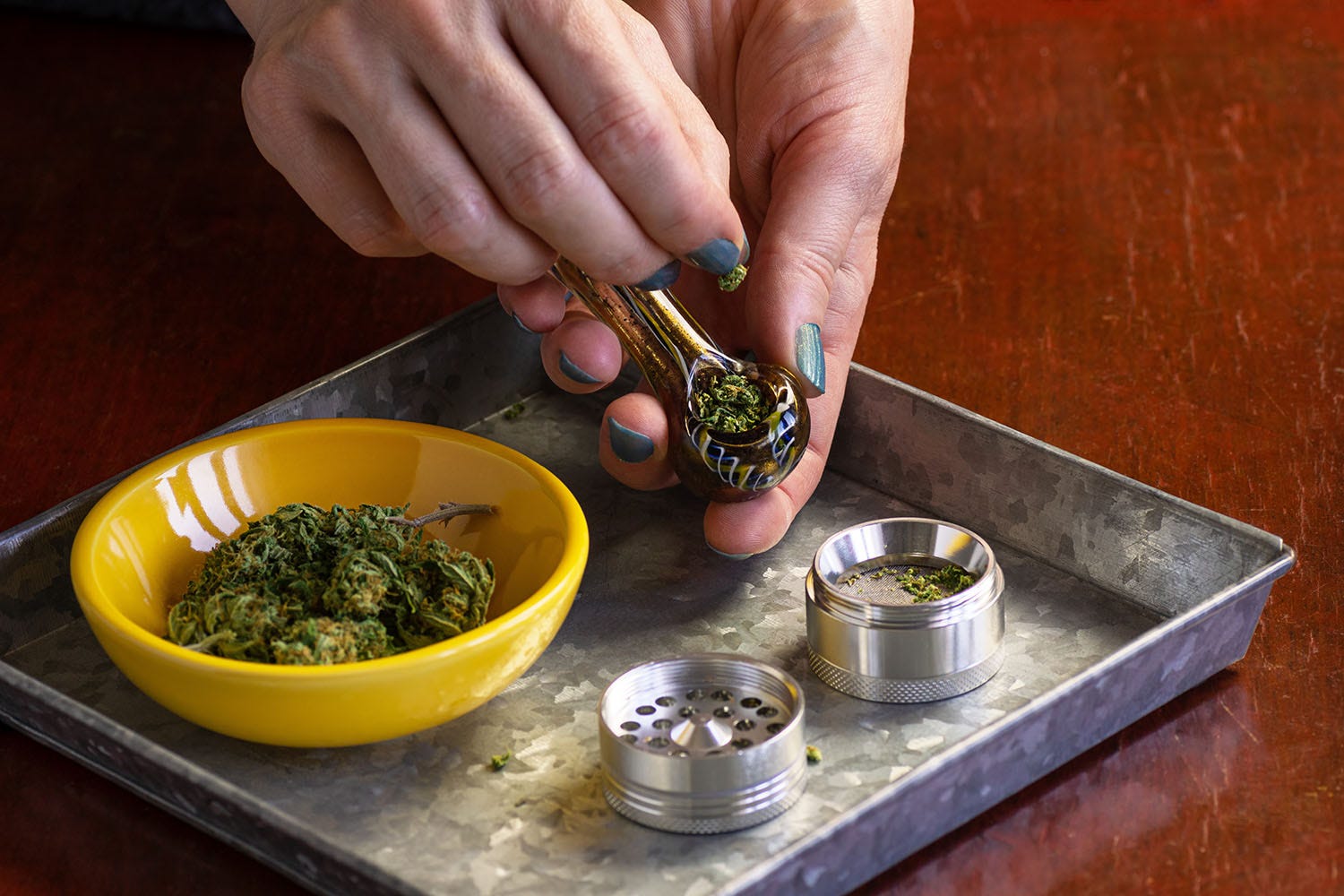 Hands adding marijuana flower to the bowl of a glass pipe, over a rolling tray