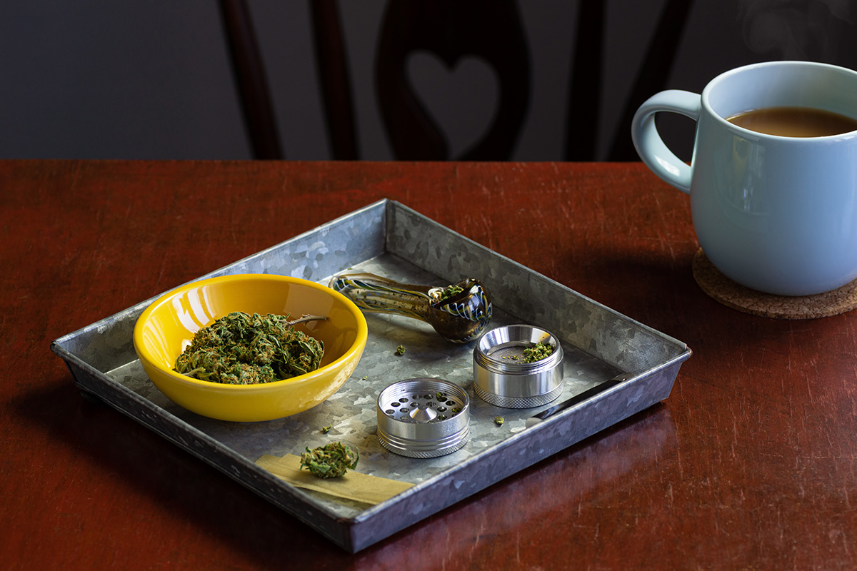 Cannabis rolling tray with flower, a grinder, and a pipe on it; next to a cup of coffee